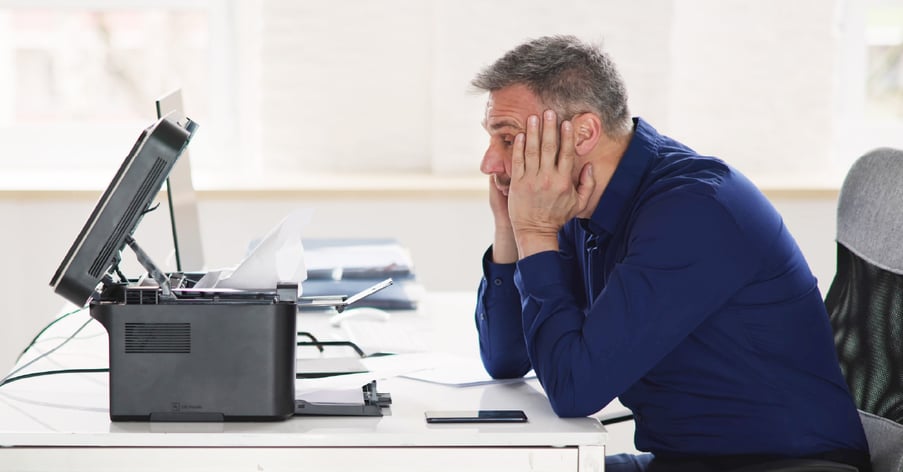 A male employee is frustrated trying to troubleshoot an A3 printer at his desk.