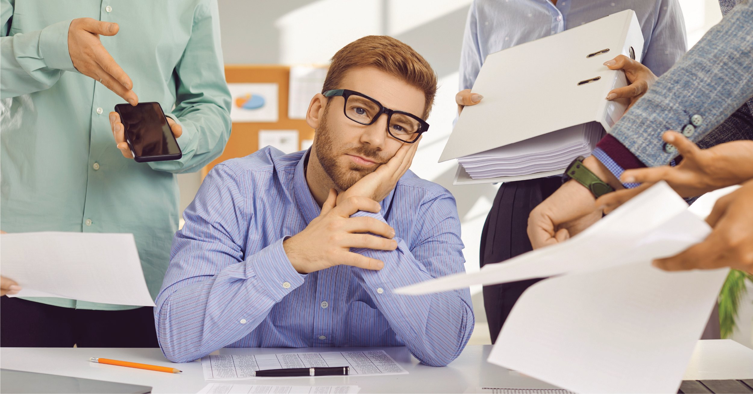 A male employee at his desk getting overwhelmed by multiple coworkers pushing additional tasks at him in the form of binders, paperwork, and another phone. 